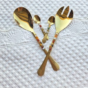 May include: A set of four gold-colored serving utensils: a spoon, a fork, and two smaller spoons. The handles are adorned with colorful beads. The utensils are arranged on a white, quilted surface.