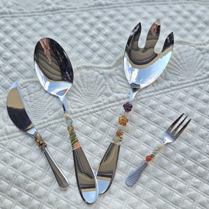 May include: A set of four stainless steel serving utensils: a knife, spoon, fork, and serving fork. Each handle is adorned with colorful beads and wire wrapping. The utensils are arranged on a white, textured surface.