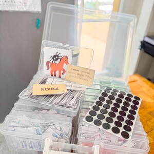 May include: Clear plastic storage boxes filled with educational resources. The boxes contain flashcards, labels with the word "NOUNS", and a sheet of black circular stickers. One card features a cartoon horse and spider.