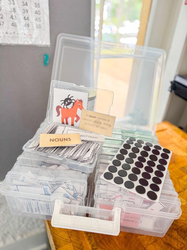 May include: Clear plastic storage boxes filled with educational resources. The boxes contain flashcards, labels with the word "NOUNS", and a sheet of black circular stickers. One card features a cartoon horse and spider.