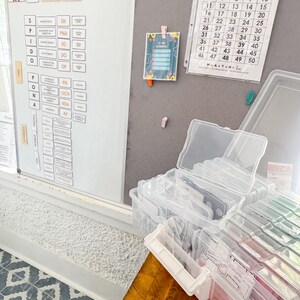 May include: A classroom setting featuring a whiteboard, a number chart, and clear plastic storage containers. The whiteboard displays grammar terms. A number chart lists numbers 1-50. The containers hold flashcards and other educational materials.