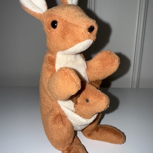 May include: A soft toy kangaroo in brown and cream colours. The kangaroo has large ears with white inner linings, black eyes, and a pouch holding a smaller joey. The toy is made of soft, textured fabric.