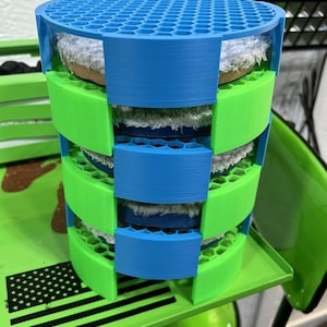 May include: A multi-tiered, cylindrical organizer in blue and green, holding several circular buffing pads. The organizer has a honeycomb pattern on the top and is made of plastic. The pads are white and tan.