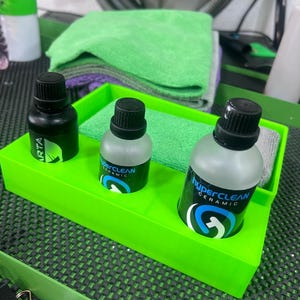 May include: A bright green plastic tray holds three bottles with black caps and labels. Two bottles are clear with "hyperCLEAN CERAMIC" labels, and one is black with a white label. Green and purple microfiber cloths are in the background.