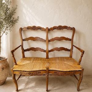 May include: A wooden bench featuring a double-back design with carved floral details on the top rails and front apron. The seat is constructed from woven rush material, supported by curved legs and armrests with a warm brown wood finish.