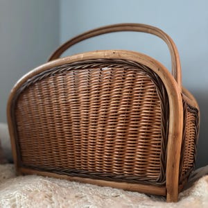 May include: A woven wicker magazine holder with a brown and tan color scheme. The holder has a curved top and a handle.
