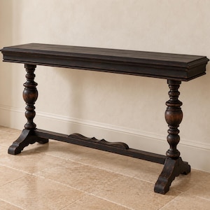 May include: A long, rectangular console table with a dark brown, distressed wood finish. The table features a flat top with molded edges and is supported by two vertical legs with turned, bulbous detailing, connected by a low, arched stretcher.