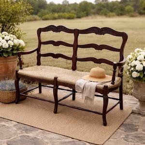 May include: A dark brown wooden bench with a woven seat and back. A straw hat and a light-colored scarf rest on the seat. The bench is outdoors, with plants and a grassy field in the background.