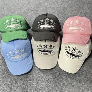 May include: A collection of six trucker caps in green, black, pink, light blue, off-white, and white. Each cap features an embroidered design on the front panel consisting of a central ship graphic surrounded by five stars in a curved arrangement.