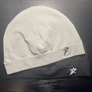 Op de afbeelding: Twee mutsen, één lichtgrijs en één zwart, zijn op een donkere ondergrond gestapeld. Elke muts heeft een klein, geborduurd sterembleem op de voorkant. De hoeden lijken gemaakt van zacht, gebreid materiaal.