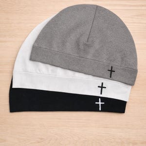 May include: Three beanies in a stack, each with a small embroidered cross. The top beanie is gray, the middle one is white, and the bottom one is black. The beanies are made of a soft material and are suitable for everyday wear.