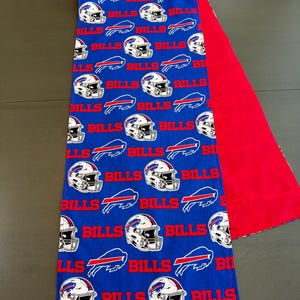 May include: A rectangular fabric table runner featuring a blue background with a repeating pattern of white football helmets, red buffalo logos, and the word BILLS in red text. The reverse side of the table runner is a solid bright red fabric.