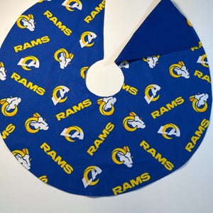 May include: A circular blue fabric tree skirt featuring a repeating pattern of the Los Angeles Rams logo and the word RAMS in yellow and white. The skirt has a central circular cutout and a single slit extending to the outer edge for placement.