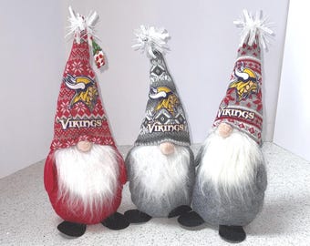 May include: Three decorative gnomes with long white beards and black shoes. Each gnome wears a knit hat with the word "Vikings" and a team logo. The hats are red, gray, and gray with white snowflake patterns and white pom-poms.