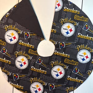 May include: A black and gray tree skirt featuring the Pittsburgh Steelers logo and team name. The skirt has a circular shape with a cutout for the tree trunk and is made of fabric with a repeating pattern.
