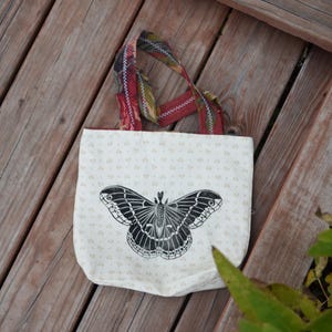 May include: Cream-colored tote bag with a large black butterfly design. The bag has a floral pattern and colorful straps. The bag is sitting on a wooden surface.