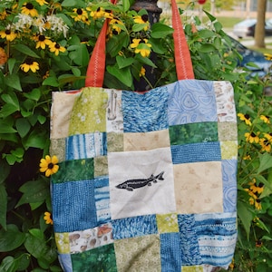 May include: A patchwork tote bag with orange handles. The bag features a variety of blue, green, and beige fabric squares. A white square displays a black illustration of a fish. The bag is hanging in front of yellow flowers and green foliage.