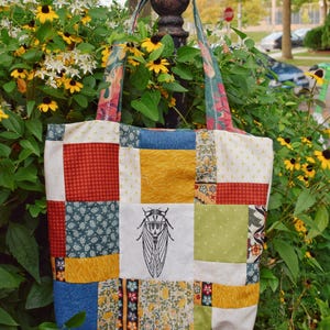 May include: A patchwork tote bag with a variety of fabric patterns in red, blue, yellow, and green. The center square features a black and white illustration of a cicada. The bag has matching patterned handles.
