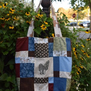 May include: A patchwork tote bag with a variety of fabric patterns in shades of blue, brown, and cream. The center panel features a black and white illustration of two morel mushrooms. The bag has matching patterned handles.
