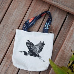 May include: A white tote bag with a black heron illustration in flight. The bag has navy blue handles with a floral pattern in orange, blue, and white. The bag is made of a light-colored fabric with small white polka dots.