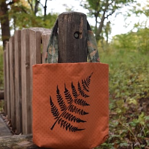May include: An orange tote bag with a black fern leaf design. The bag has patterned handles with a green and brown floral print. The bag is hanging on a wooden post in a natural outdoor setting.