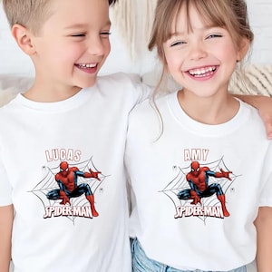 Könnte beinhalten: Weiße T-Shirts mit einem Spider-Man-Motiv und den Namen "LUCAS" und "AMY" über dem Bild. Das Spider-Man-Design zeigt einen rot-blauen Superhelden in einem Netz. Die T-Shirts werden von zwei lächelnden Kindern getragen.