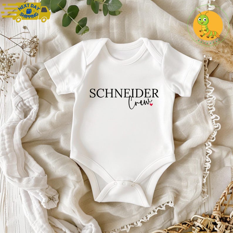 May include: White baby onesie with the text "SCHNEIDER Crew" in black script. A small red heart is next to the word "Crew". The onesie is short-sleeved and has a snap closure at the bottom. The onesie is laying on a beige cloth.
