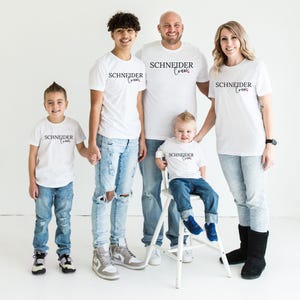 May include: A family of five wearing matching white t-shirts with the text "SCHNEIDER Crew" in black script. The family is standing in front of a white background. The adults and older children are wearing blue jeans.