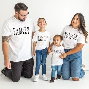 May include: Four people wearing white t-shirts with the word "FAMILY" printed in black. The shirts also include names and hearts. The people are wearing blue jeans and black trousers. The family is smiling and posing for a photo.