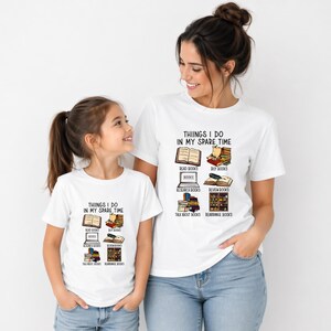 Op de afbeelding: Twee witte t-shirts met de tekst "THINGS I DO IN MY SPARE TIME" en illustraties van boeken. De shirts worden gedragen door een vrouw en een kind. De illustraties tonen open boeken, stapels boeken en een boekenplank.