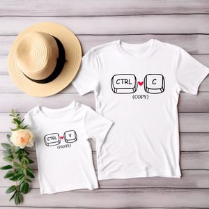 Op de afbeelding: Twee witte T-shirts met zwarte toetsenbordafbeeldingen. Eén shirt toont "CTRL" en "C" met een hart, met het label "(COPY)". Het andere shirt toont "CTRL" en "V" met een hart, met het label "(PASTE)". Een strohoed en een bloemstuk staan ook in de afbeelding.