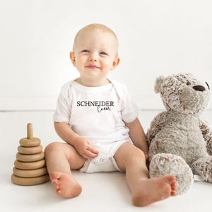 May include: A smiling baby wearing a white onesie with the text "SCHNEIDER Crew" in black script. The baby is sitting next to a wooden stacking toy and a gray teddy bear on a white surface.