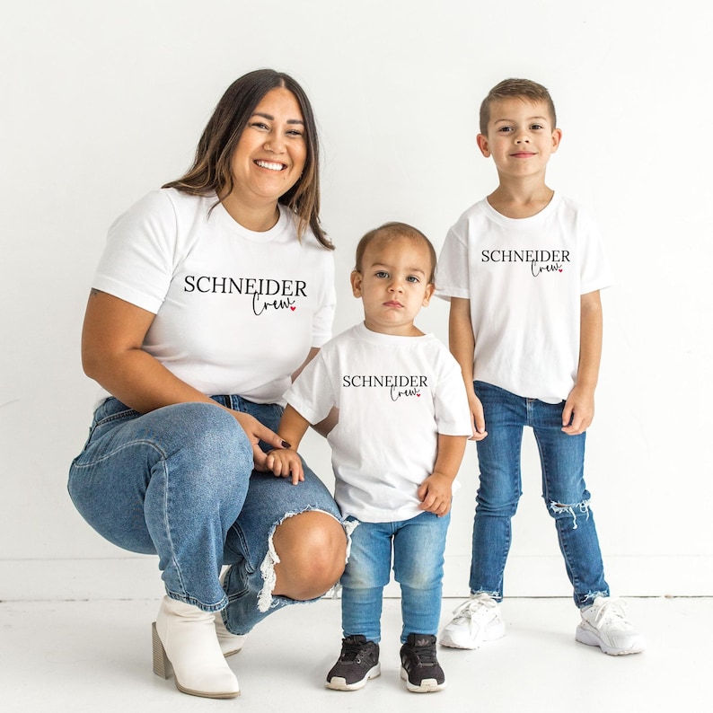 Könnte beinhalten: Drei Personen tragen weiße T-Shirts und blaue Jeans. Die Shirts tragen den schwarzen Aufdruck "SCHNEIDER Crew". Die Person links kniet, die anderen beiden stehen. Der Hintergrund ist schlicht weiß.