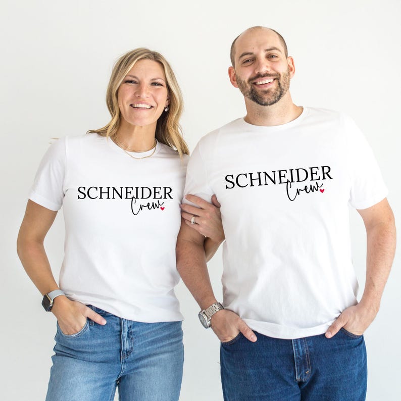 May include: Two people wearing white t-shirts with the text "SCHNEIDER Crew" in black script. The word "Crew" has a heart symbol. The people are wearing blue jeans and smiling. The background is plain white.