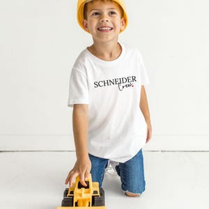 May include: A white t-shirt with the text "SCHNEIDER Crew" in black script and a small pink heart. The shirt is worn by a child wearing a yellow construction hat and blue jeans. A yellow toy construction vehicle is in the foreground.