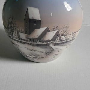 Könnte beinhalten: Keramikvase mit hellblauem Farbverlauf und Winterlandschaft. Die Szene zeigt schneebedeckte Gebäude und einen Kirchturm, mit kahlen Bäumen und einem verschneiten Weg. Die Vase hat einen breiten, abgerundeten Körper und einen schmalen Hals.