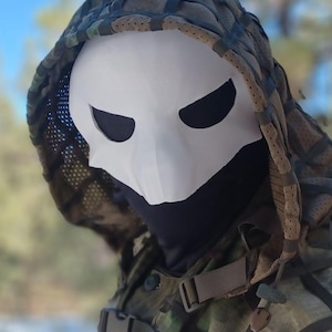 Op de afbeelding: Een wit masker met zwarte ooggaten, gedragen met een camouflagekap en jas. Het masker heeft een schedelachtig uiterlijk. De jas heeft riemen en gespen. De achtergrond is wazig, met de focus op het masker en de kleding.