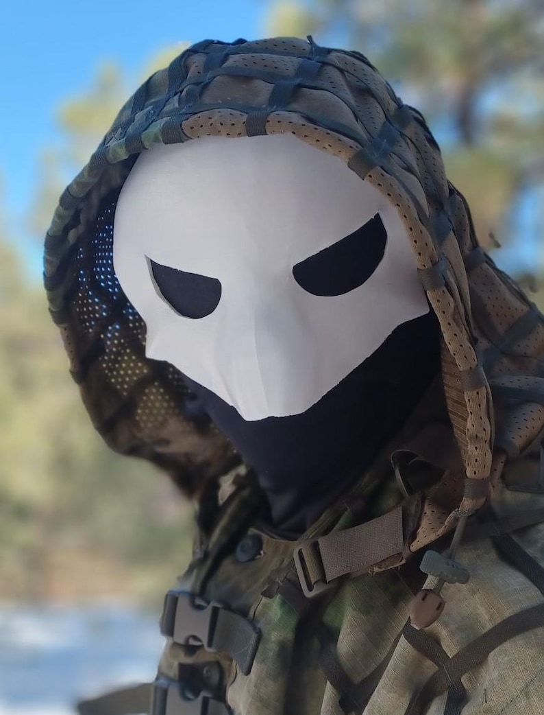 Op de afbeelding: Een wit masker met zwarte ooggaten, gedragen met een camouflagekap en jas. Het masker heeft een schedelachtig uiterlijk. De jas heeft riemen en gespen. De achtergrond is wazig, met de focus op het masker en de kleding.