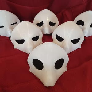 Op de afbeelding: Zes witte maskers met zwarte oogopeningen zijn gerangschikt op een rode ondergrond. De maskers hebben een gladde, afgeronde vorm, met verschillende oogontwerpen. De maskers zijn waarschijnlijk voor kostuum- of theatrale doeleinden.
