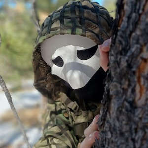 Op de afbeelding: Een wit masker met grote ooggaten wordt gedragen met een camouflagekap en -jas. Het masker heeft een spookachtige uitstraling. De persoon is gedeeltelijk verborgen achter een boom. Geschikt voor Halloween of kostuumevenementen.