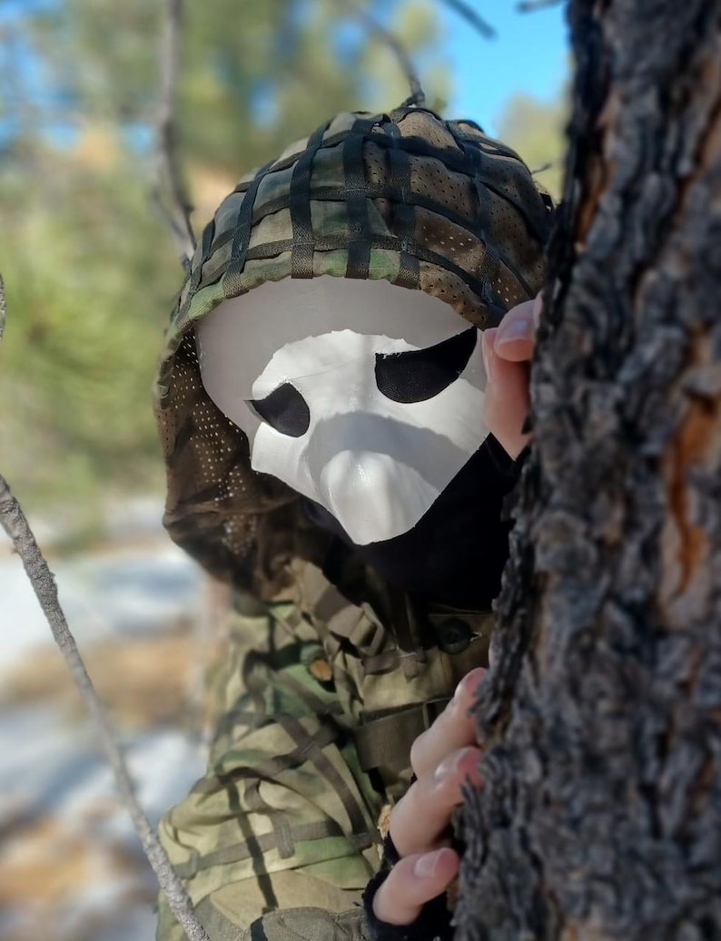 Op de afbeelding: Een wit masker met grote ooggaten wordt gedragen met een camouflagekap en -jas. Het masker heeft een spookachtige uitstraling. De persoon is gedeeltelijk verborgen achter een boom. Geschikt voor Halloween of kostuumevenementen.