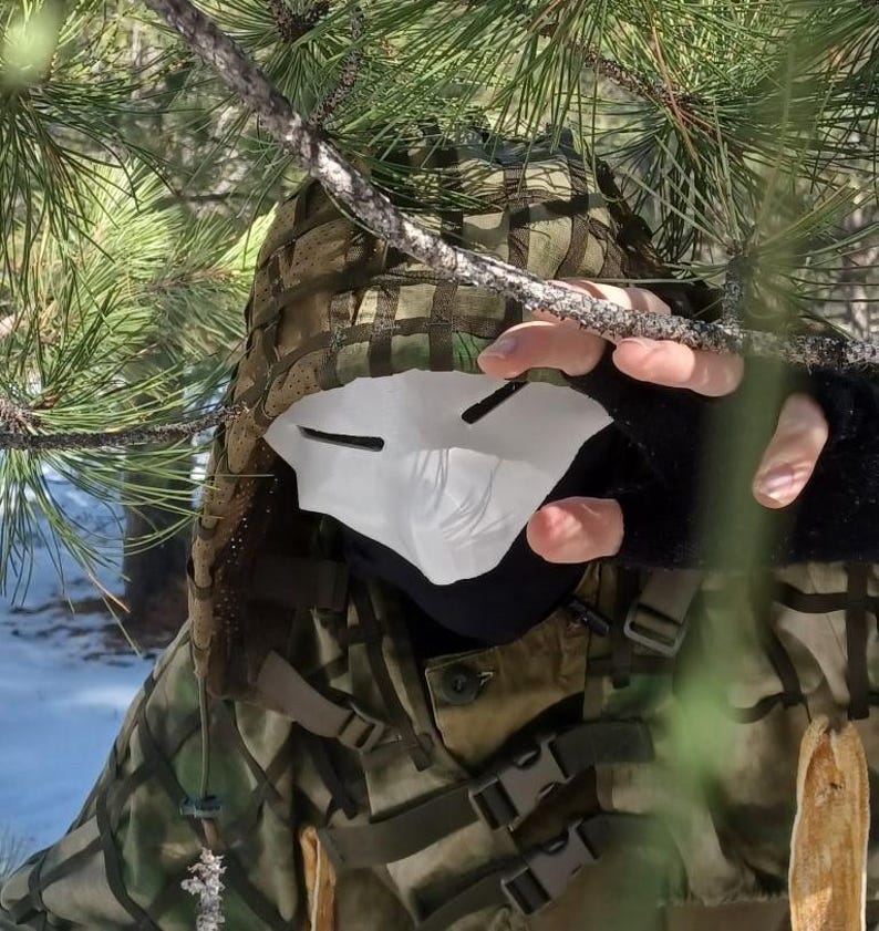 Op de afbeelding: Een camouflagejas en een wit masker met zwarte oogspleten. De persoon wordt gedeeltelijk verborgen door dennentakken, wat een gevoel van mysterie en verberging cre&euml;ert. De jas heeft meerdere gespen.