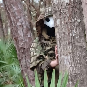 Op de afbeelding: Een persoon die een camouflage jack met capuchon en een wit masker met zwarte ooggaten draagt, gedeeltelijk verborgen door bomen en bladeren. De persoon bevindt zich in een bosrijk gebied met groene planten en bruine boomstammen.