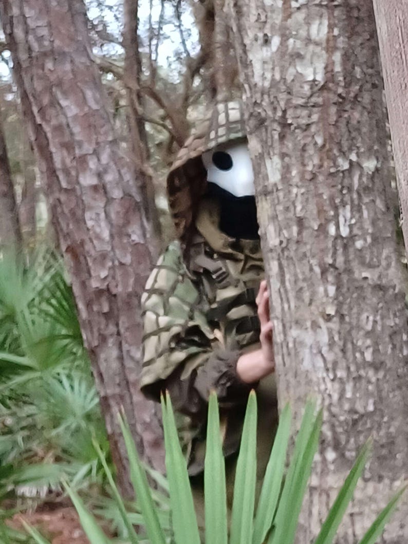 Op de afbeelding: Een persoon die een camouflage jack met capuchon en een wit masker met zwarte ooggaten draagt, gedeeltelijk verborgen door bomen en bladeren. De persoon bevindt zich in een bosrijk gebied met groene planten en bruine boomstammen.