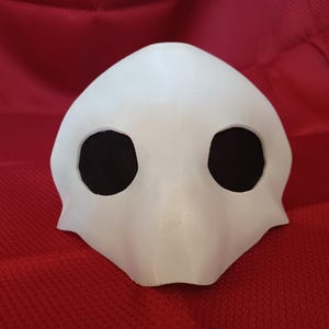Op de afbeelding: Een wit masker met grote, ovale ooggaten en een snavelachtige uitsteeksel. Het masker is gemaakt van een glad, stevig materiaal en staat tegen een rode achtergrond. Het ontwerp van het masker suggereert een kostuum of theatrale prop.