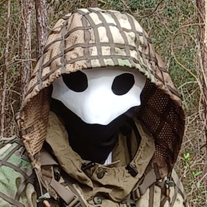 Op de afbeelding: Een persoon die een wit pestdoktermasker draagt met zwarte ooggaten, een zwarte nekbedekking en een camouflage capuchonjas. De capuchon is bedekt met een bruin en tan net.