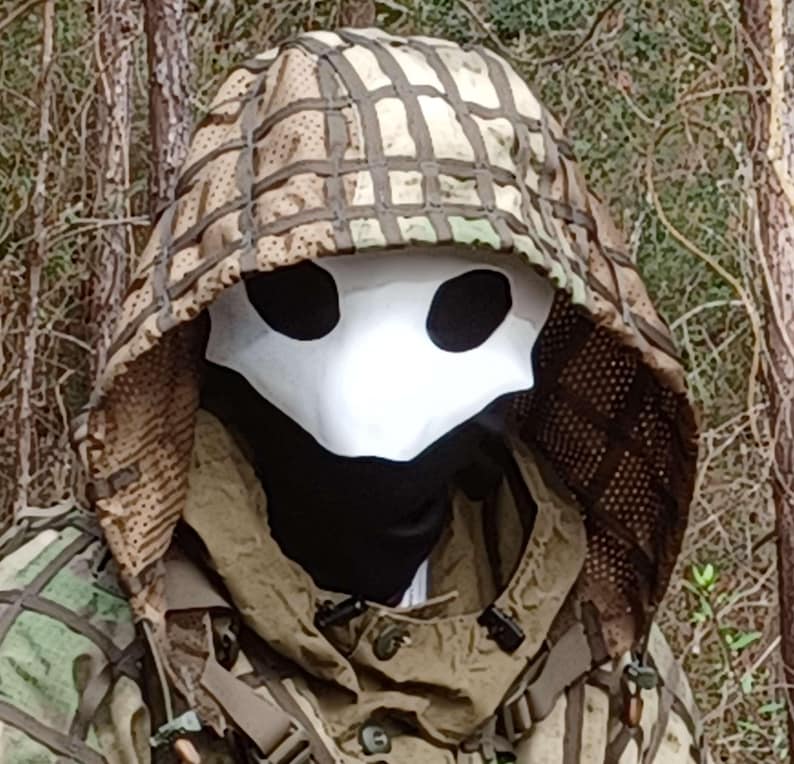 Op de afbeelding: Een persoon die een wit pestdoktermasker draagt met zwarte ooggaten, een zwarte nekbedekking en een camouflage capuchonjas. De capuchon is bedekt met een bruin en tan net.