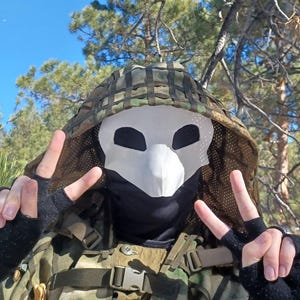 Op de afbeelding: Een persoon draagt een wit masker met grote oogopeningen en een snavelachtige neus, gecombineerd met een camouflagekap en -jas. De persoon maakt een vredesteken met zijn vingers. De achtergrond toont bomen en een blauwe lucht.
