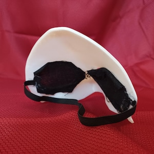 Op de afbeelding: Een wit masker met zwarte oogbedekkingen en een zwarte elastische band. Het masker ligt op een rode ondergrond. Het masker is waarschijnlijk voor kostuum- of theatrale doeleinden.