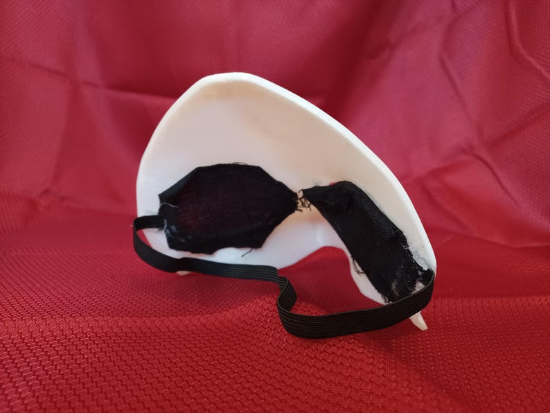 Op de afbeelding: Een wit masker met zwarte oogbedekkingen en een zwarte elastische band. Het masker ligt op een rode ondergrond. Het masker is waarschijnlijk voor kostuum- of theatrale doeleinden.